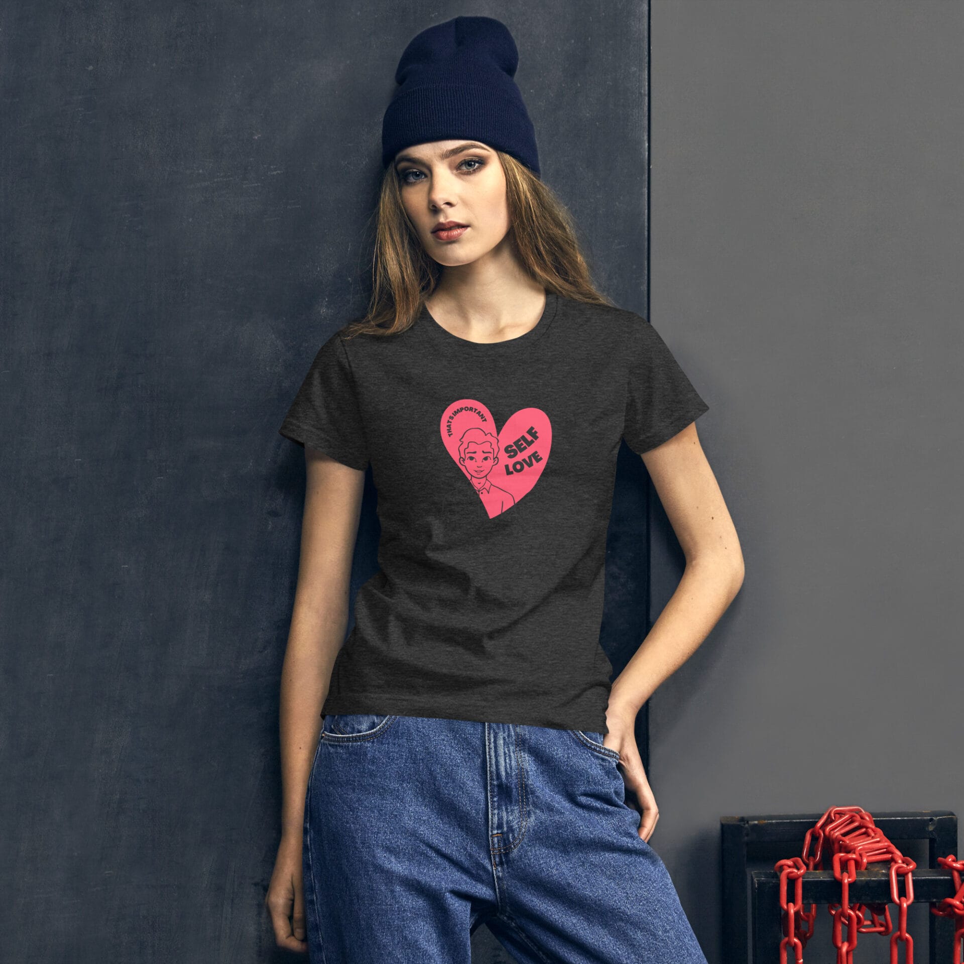 A woman poses wearing a dark t-shirt with a pink heart graphic featuring the text "SELF LOVE" on it paired with casual denim jeans against a textured gray background.