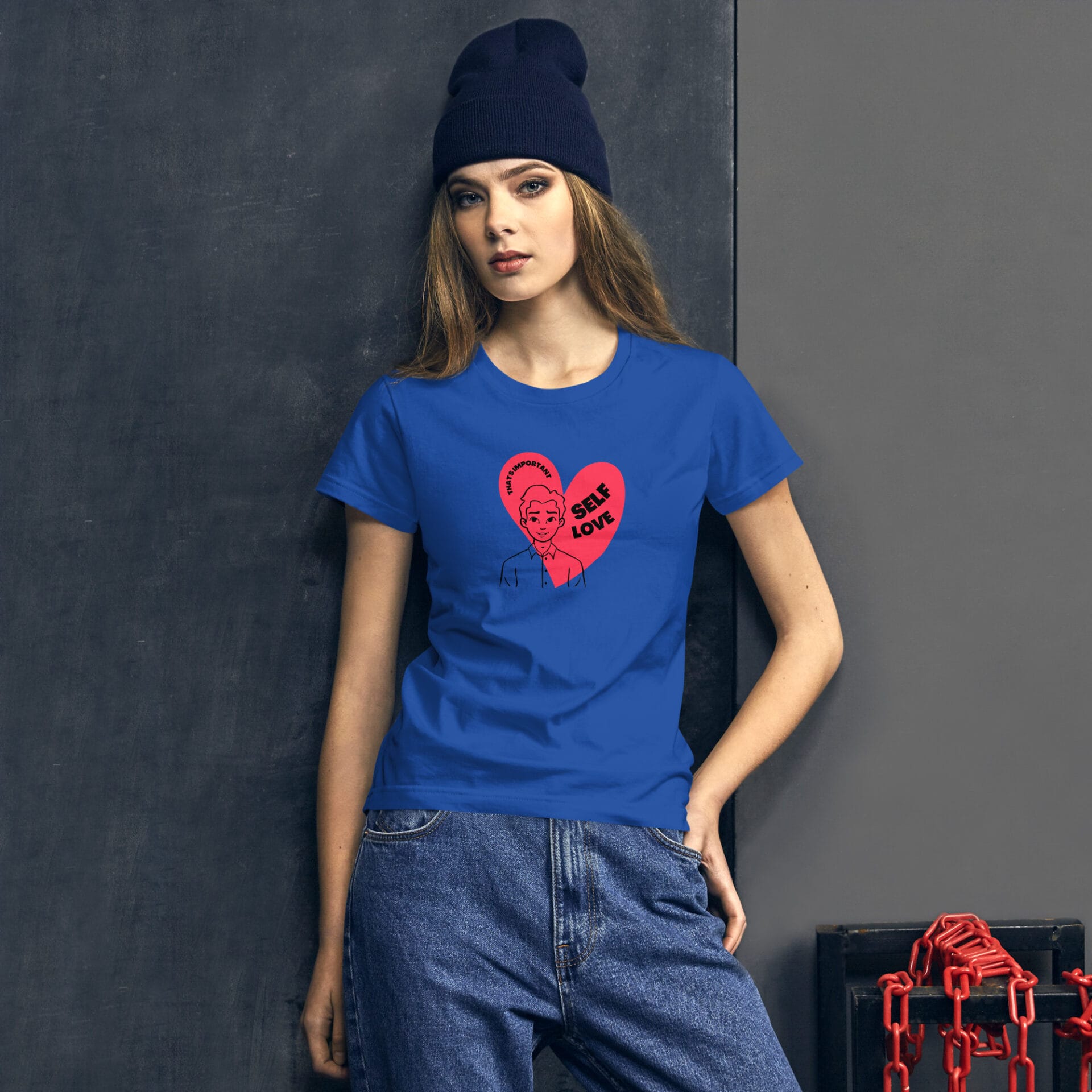 A young woman wearing a blue t-shirt with a graphic design stands confidently against a dark wall. Her outfit is complemented by a dark beanie and she appears relaxed.