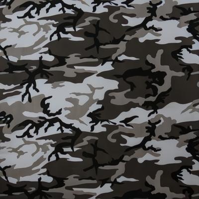 A fabric patterned with various shades of green black and white resembling a camouflage design is laid flat against a neutral background indicating potential use in clothing or outdoor gear.