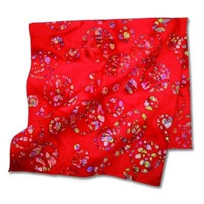A red fabric square features vibrant circular patterns with contrasting colors. It lies flat  showcasing its texture and design in a bright  light setting.