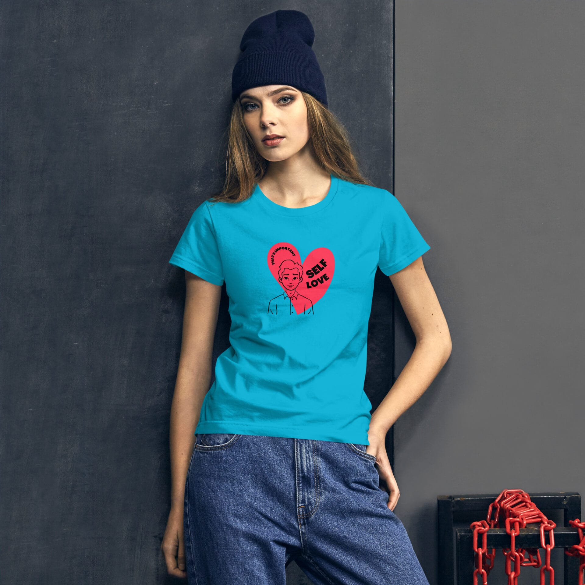 A young woman stands confidently in a bright blue t-shirt featuring a graphic of a cartoon figure and the text "SELF LOVE" within a red heart design against a dark background.