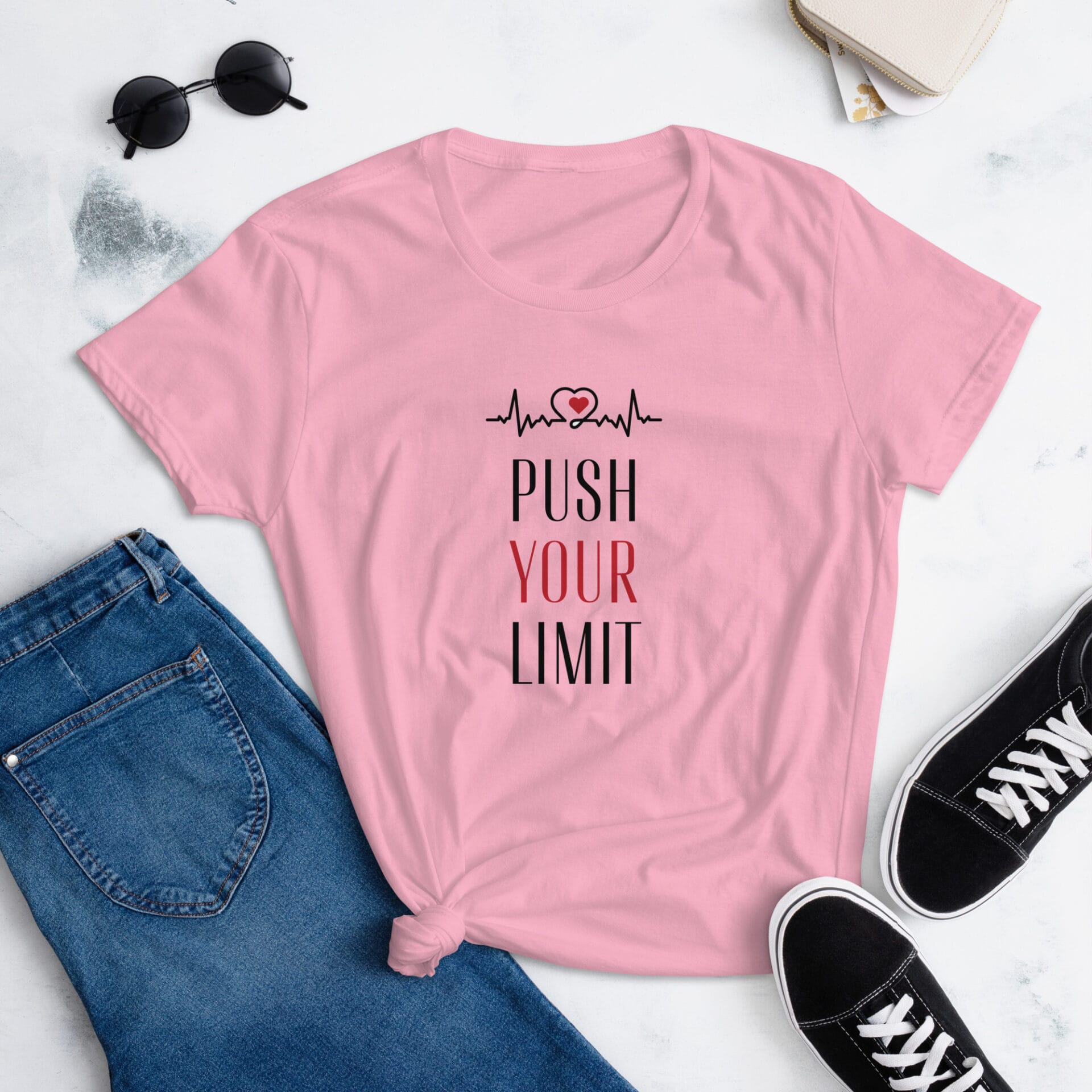 A pink t-shirt with the text PUSH YOUR LIMIT and a heart with a heartbeat line is displayed alongside denim jeans and black sneakers in a light  neutral setting.