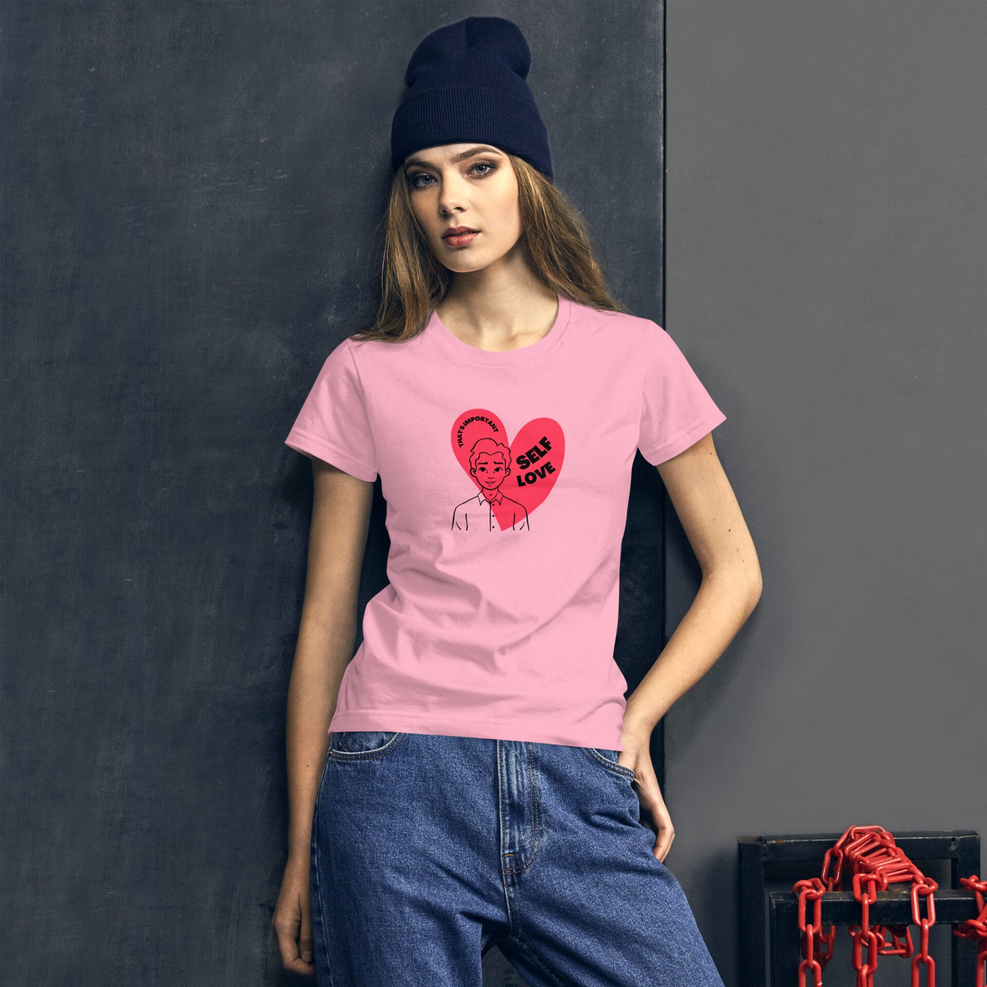 A woman stands against a dark wall wearing a pink t-shirt with an illustration of a person and the text "SELF LOVE" in a heart  paired with blue jeans and a beanie.
