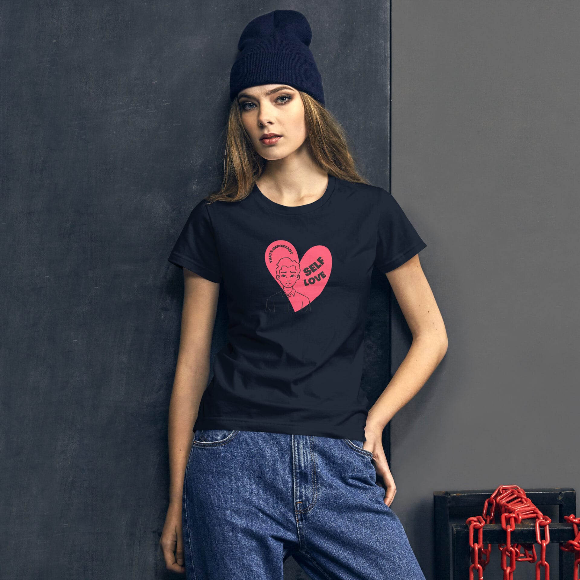 A young woman wears a black t-shirt featuring a pink heart graphic with the text "SELF LOVE" and a figure. She stands against a dark wall with a decorative chain nearby.